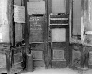 A black and white photograph depicts the rustic wooden entrance of a workshop. Several signs hang on the building's edifice.