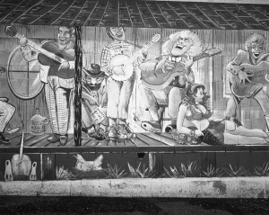 The outer wall of a building is covered with a mural of musicians. A depiction of a scantily clad woman in a relining pose occupies the lower right corner of the mural. At the base of the mural are images of plants, a chicken, and the backside of a burrowing dog.