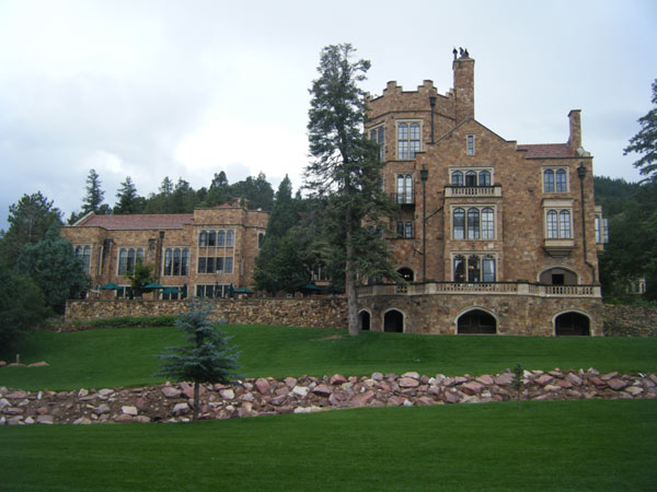 A photo of the castle and carriage house on the grounds with vibrant green grass and pine trees throughout the picture. 