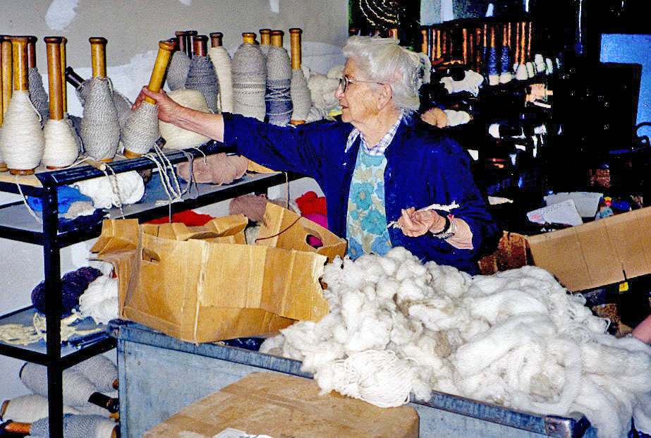 Image of an older woman, standing next to a large bin full of loose white wool fibers and shelves upon which stand many spindles of different colors of wool. She is looking at the spindles and with her right hand she is selecting a spindle that is half full of grey wool.