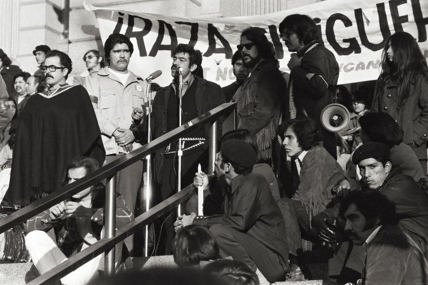 Photo of an outdoor anti-war protest rally featuring speaker Corky Gonzales. He stands in front of a microphone flanked by unidentified men and women standing and sitting on the steps. A banner behind Corky reads "RAZA SI, GUERRA NO."