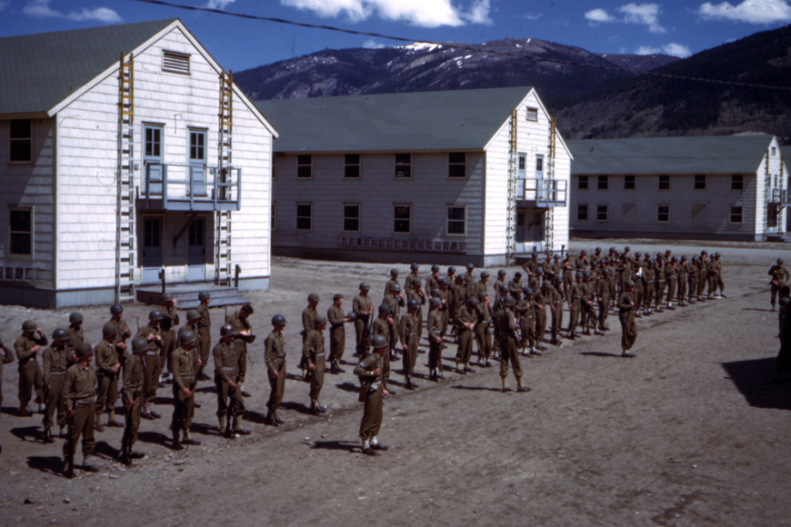 A large group of soldiers at Camp Hale, standing in formation in front of 3 large, white barracks. They wear uniforms and helmets. In the background you can see the mountains and a blue sky.