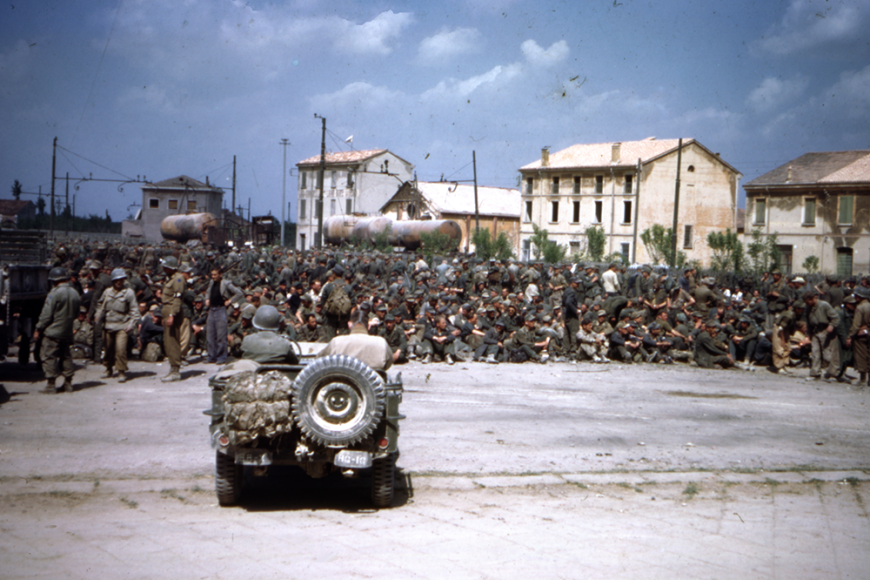 A very large group of soldiers sits in the center of Camp Hale with an army vehicle out front. There are white buildings, sparse trees, and gas tanks in the background.