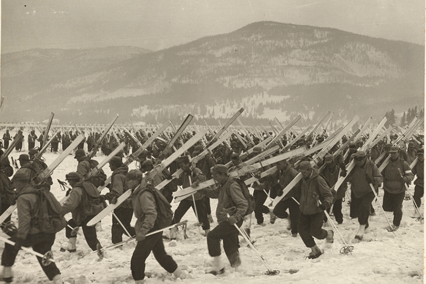 A large group of soldiers are trudging across a snowbank in Camp Hale. They each hold a pair of skis and poles.