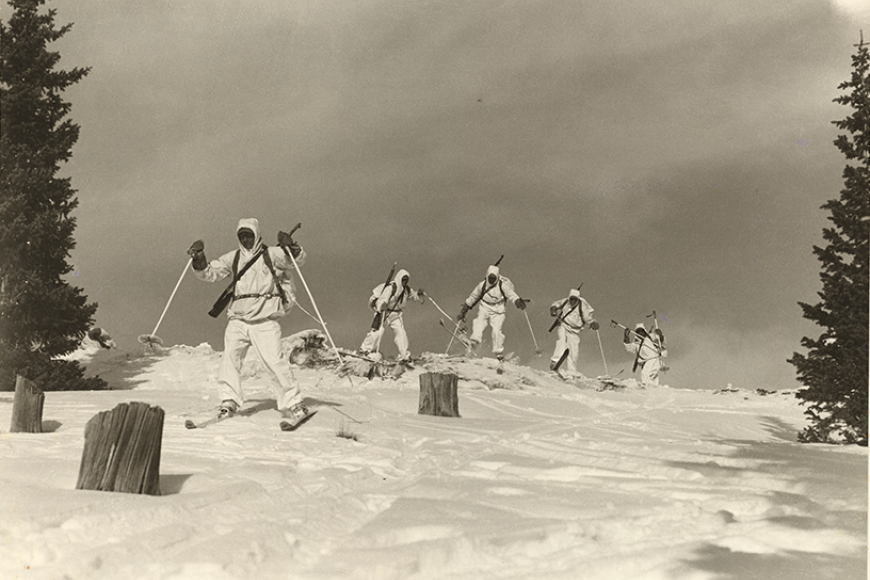 A black and white photo of 5 soldiers in white snow gear skiing with poles down a slope. There are 3 logs placed in front of them as obstacles. One soldier is jumping in the air.