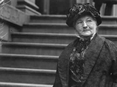 Black and white photograph of Mary Harris "Mother" Jones standing in a coat on the entry stairs of a building.