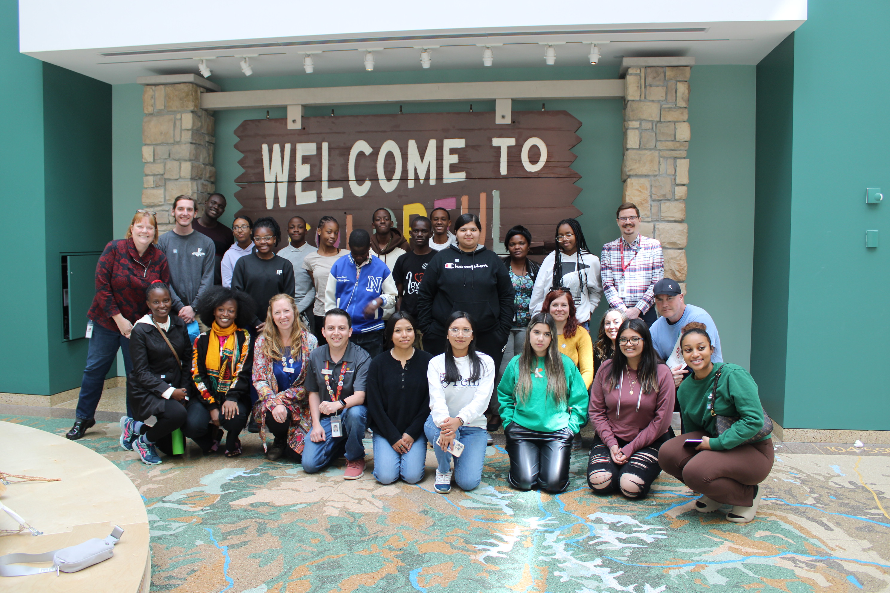 A large group of students posed with various History Colorado staff in the History Colorado Center Lobby in front of a large wooden sign reading "Welcome to Colorful Colorado"