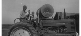 A man and two children pose on a John Deere tractor in a black and white photograph dating to 1955..