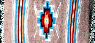 Photo of a woven piece by Eppie Archuleta called "Rain Cloud." The piece features a tan background, with horizontal thin stripes clustered together into two wide horizontal bands on each side of the image. These stripes are white, turquoise, red, and black in color. The center image is a blocky geometric shape of turquoise, black, red, and tan, with white feather-like edging. There is a very small turquoise dot in the very center.