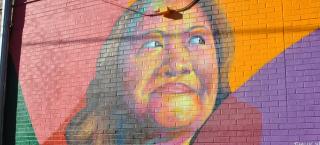 Photo of Nga Vương-Sandoval mural by Artist/Muralist Thomas Evans (I Am Detour).  The mural is an exterior brick wall, and the close up image of her face has been created using bright colors of red, orange, and pink.