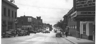 Photo of the downtown area of a smaller town, taken around the early 1940s. The street is wide, and cars are driving down the road while some are parked along both sides of the streets. Signs on one building to the left of the image says "Drugs" and "Prescriptions" while the upper floor sign says "Business College." To the right of the image, there is a theater in the foreground, advertising a matinee at 2pm and the films playing  include "Desperadoes" and "Rhythm of the Islands."