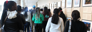 A large group of students seen from behind as they walk through a mezzanine exhibit at the History Colorado Exhibit. Framed photos and panels line the wall showing the Corn Mothers exhibition.