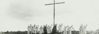 Members of the Ku Klux Klan dressed in white and black robes make a raised-arm salute before a tall cross on a rocky mountaintop