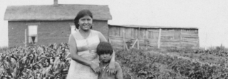 A Ute woman and young boy standing in a crop field with a wooden structure behind them.a