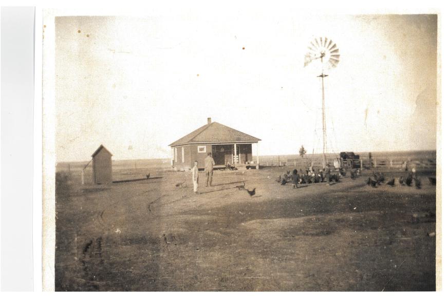 A sepia tone photograph of a man and child standing with chickens in front of a farmhouse and windmill.