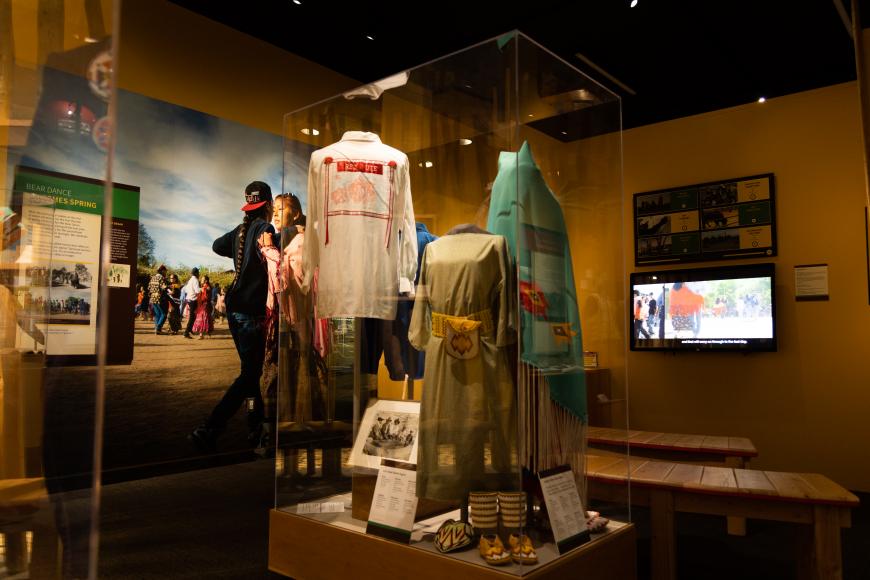 An exhibit space at Ute Indian Museum. On the walls are panels featuring landscapes and labels. In the center of the space are traditional clothing in glass cases. On one wall is a television screen providing additional information about the exhibit.