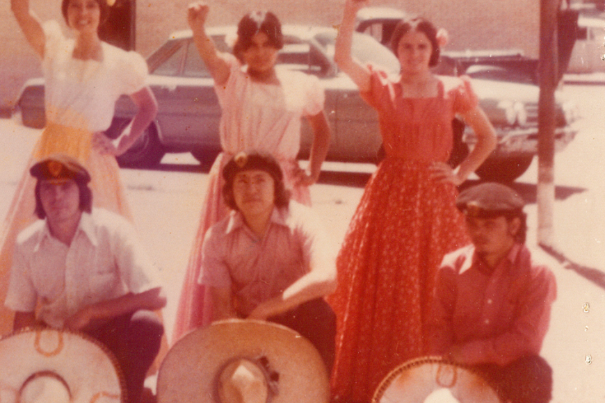 A Folklórico group from Pueblo, CO demonstrating during the 1960s. The men sit on the ground with their hats off. The young women have their right fists raised in the air.