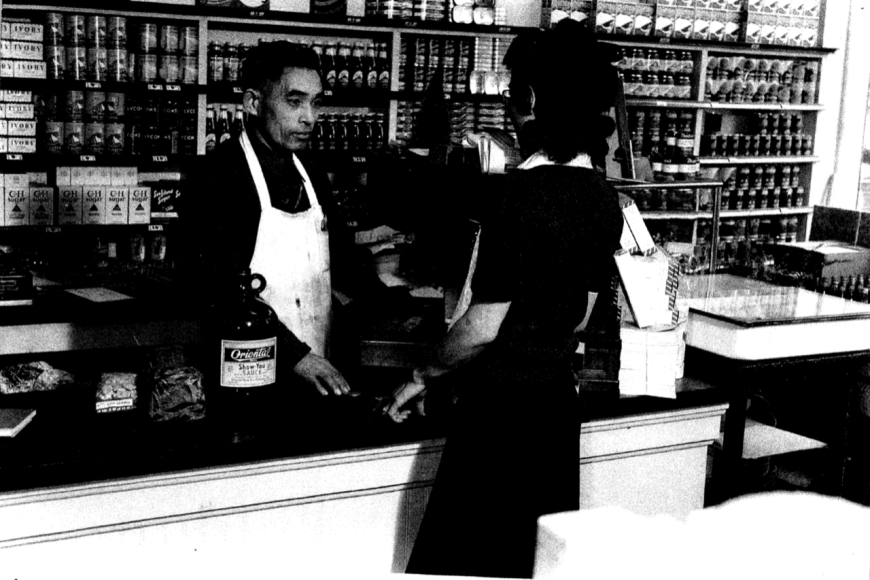 An Asian-American man stands behind the counter of a store. He wears a white apron and is speaking to a customer.