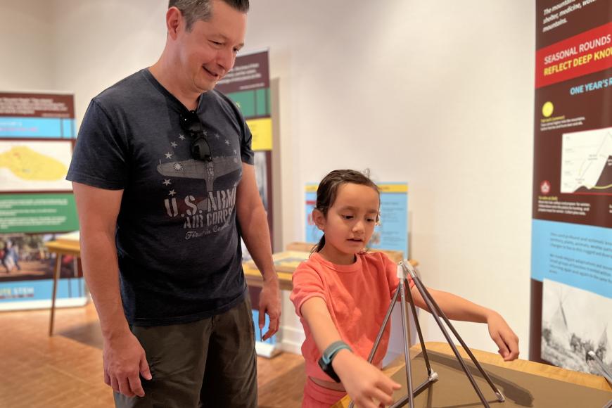 A father stands and watches beside his young daughter while she plays with a Ute Wickiup interactive piece.