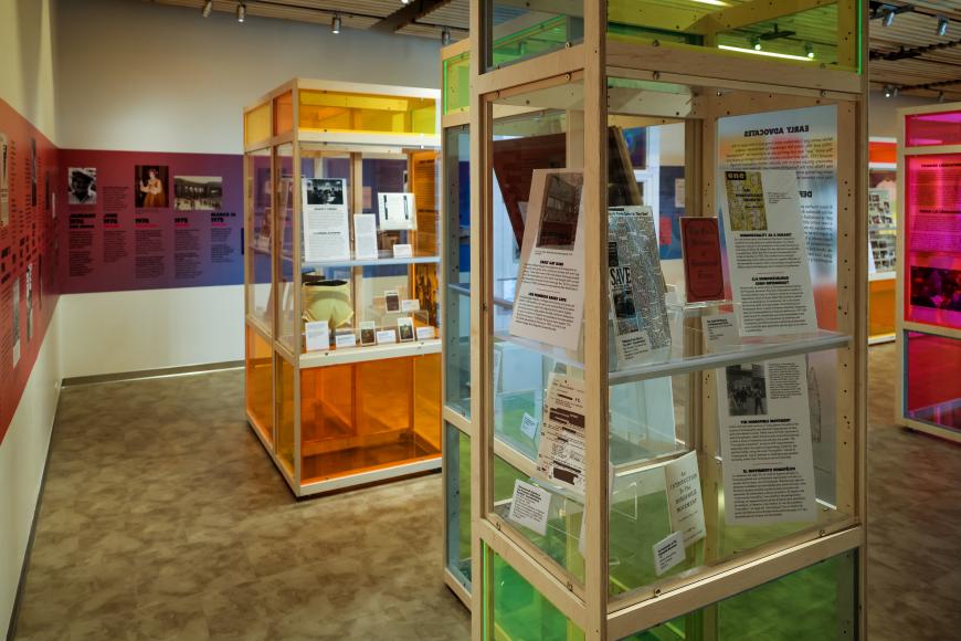 Rows of multicolored glass cases filled with artifacts and information panels, making up the Rainbows & Revolutions exhibit. There's a purple and blue timeline of LGBT history across the wall.