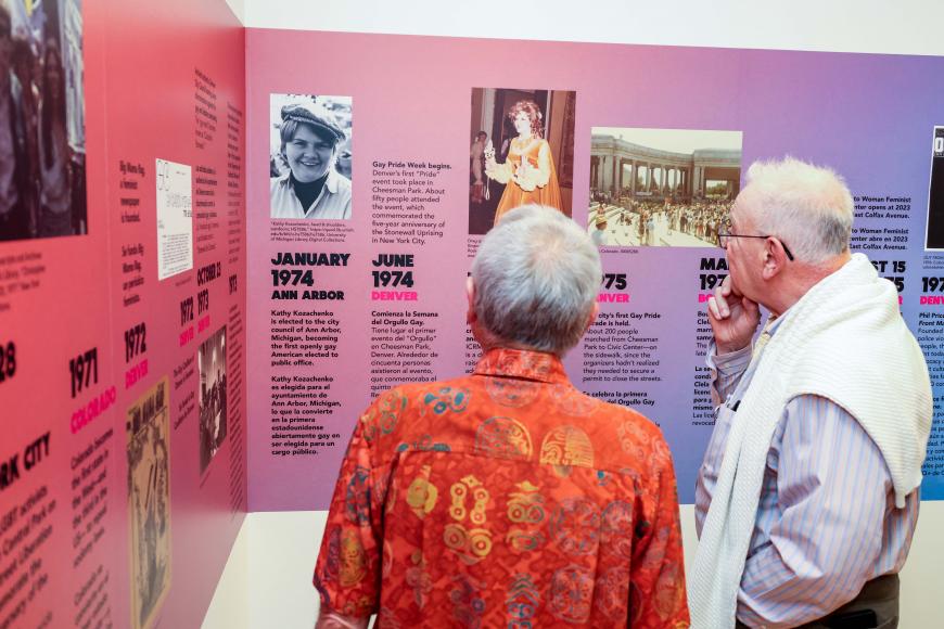 2 older gentlemen look at a purple and pink wall timeline detailing LGBT history with photos and text.