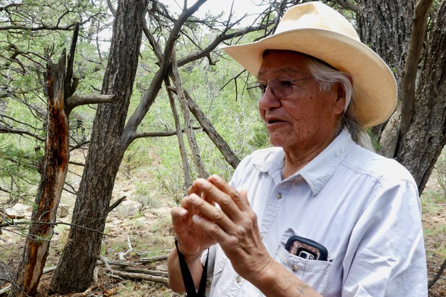 “ A male elder places his finger tips together from the Ute Mountain Ute Tribe while standing in nature speaking to a group of youth students.”