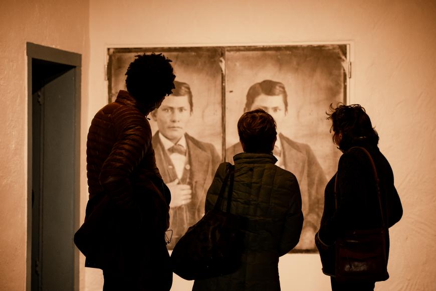 The silhouettes of 3 individuals looking at a historical portrait hung on the walls of Fort Garland. It is a young indigenous man.