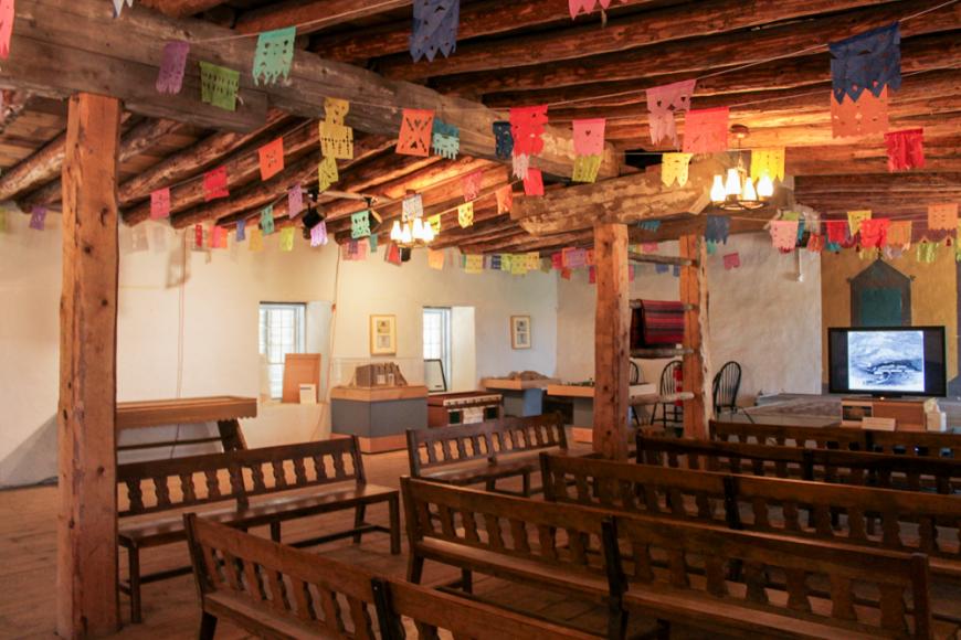 Interior of Fort Garland museum filled with benches, a tv monitor, and hanging papel picado 