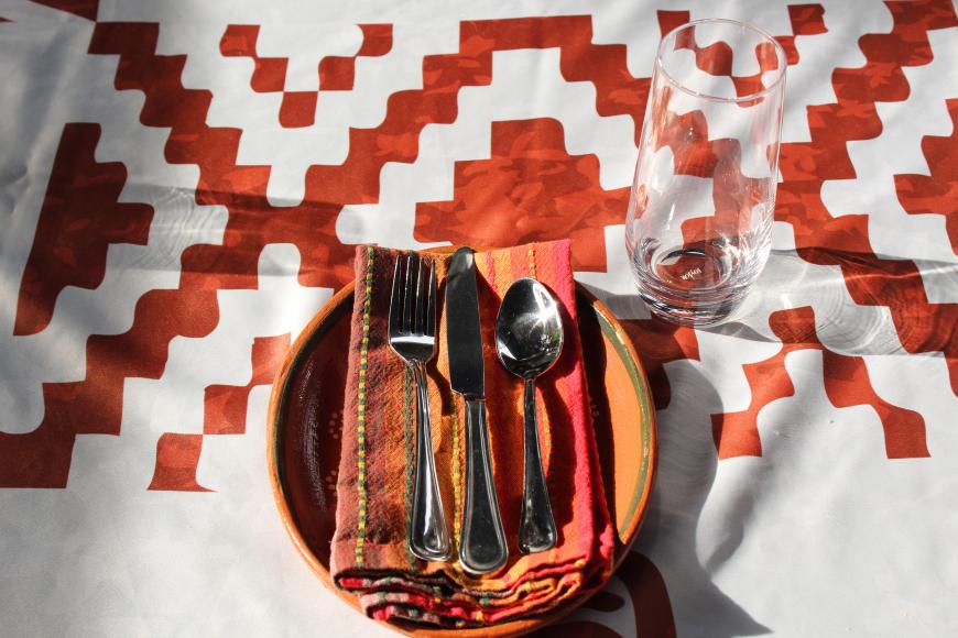 A table setting with bowl, napkin, and spoon on a mud red decorated table cloth. The napkin is vibrant red and orange.