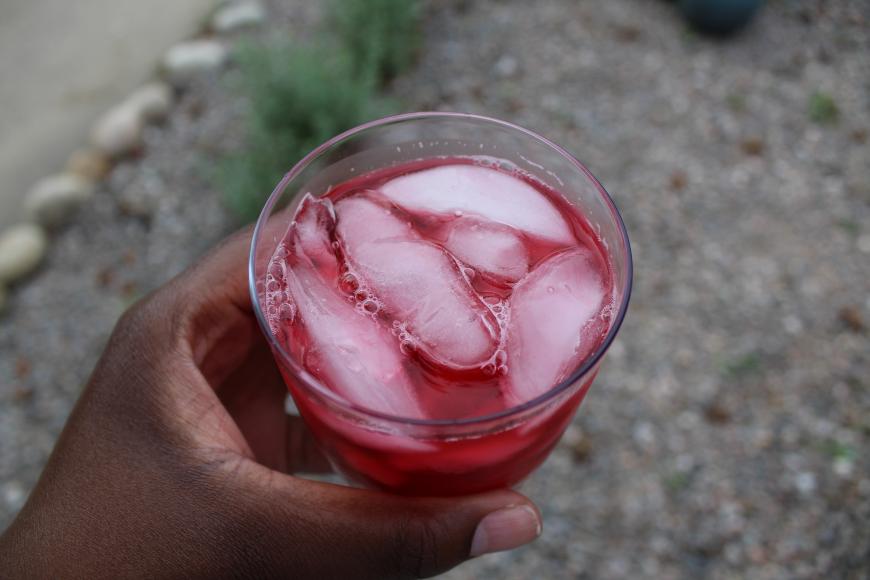An aerial view of a hand holding a glass filled with ice cubes and a bright pink drink.