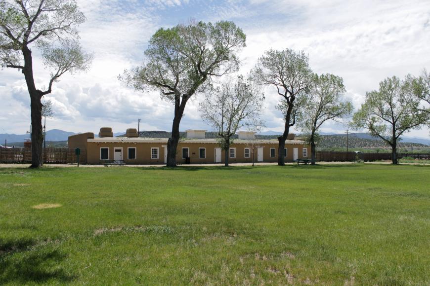 Large open green space inside Fort Garland surrounded by tall trees. The adobe buildings of the fort sit in the background.