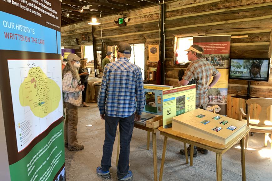 Three museum visitors engage in conversation in the middle of the Utem Stem Traveling Exhibit. Displayed around them are two pop-up banners and interactive tables, and one touch screen monitor playing an educational video.