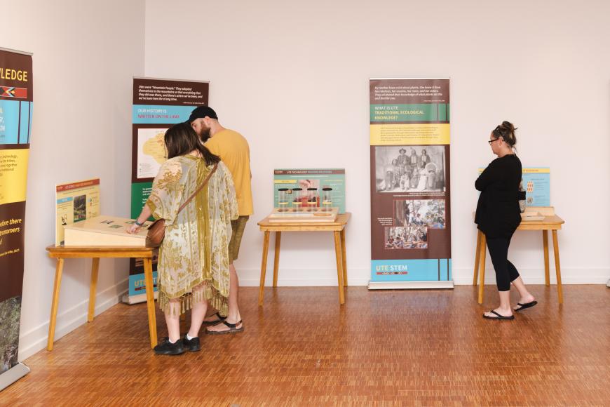 Three people walk around a museum in a room featuring the Ute Traveling Exhibit, while engaging in interactives and looking at educational pop-up banners.