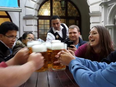 Photo of a group of friends gathered around a long wooden table at a bar or restaurant. The are each holding up a large mug of beer to the center of the table and offering a toast. There is a server at the end of the table, picking something up from the tabletop although it is obscured by the mugs of beer in the forefront. Behind the table of friends, white stone walls of the building are seen, as well as an arched doorway, and other patrons. 