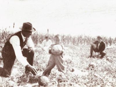 A Black family, including two adult men and a small child kneel in a field. The child holds a large vegetable and is smiling for the camera.