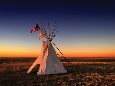 A tipi at sunset. Over the tipi fly two flags: the flag of the United States, and the white flag of peace.