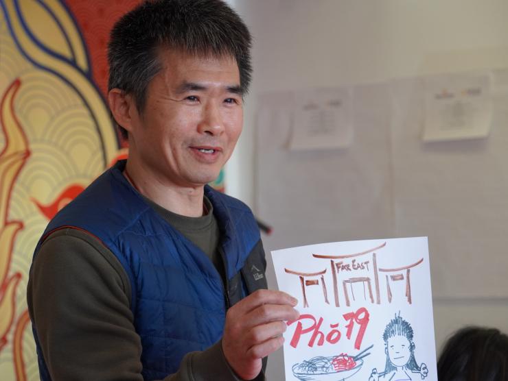 An adult man at the Little Saigon memory workshop holds a colorful hand drawn photo depicting the sign to Pho 79, the Far East Center's red, chinese archiecture-inspired gates and a simple drawing of a person with black hair.