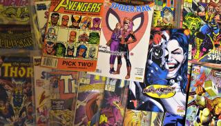 Photo of a selection of various comic books, laid out on a flat surface so that we can see some or all of each cover. Some of the comic books appear to be older and some are newer, and a few of them are inside of a clear plastic storage sleeve.