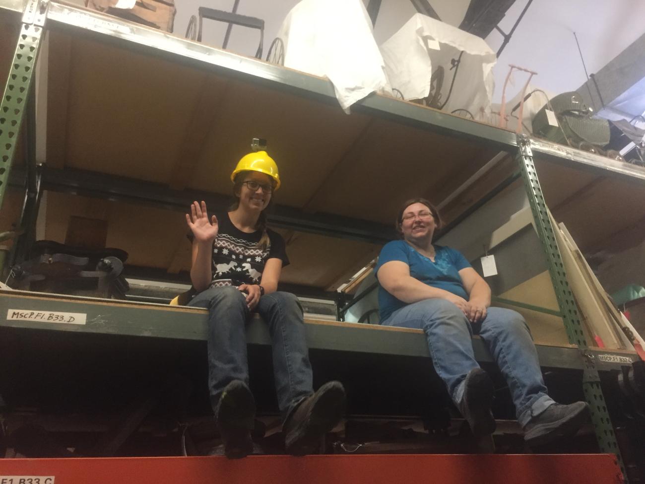 Melissa de Bie and another History Colorado employee sit on shelves in a warehouse they were emptying. They are staring in the camera with friendly smiles.