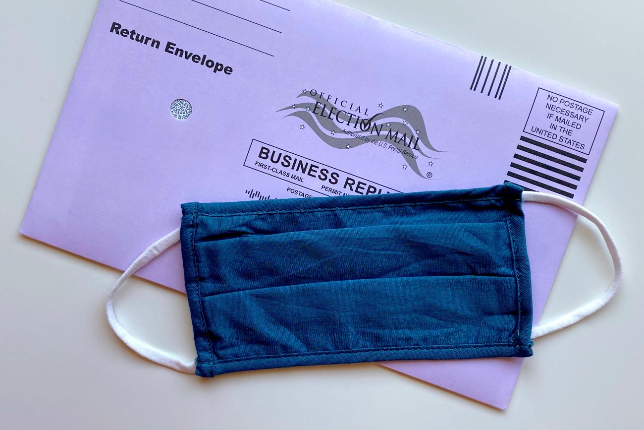 Photo of a purple mailing envelope for mailing in an election ballot. In front of the envelope, where the address is printed, is a blue demin homemade face mask used during the Covid-19 pandemic to cast their votes in the 2020 elections.