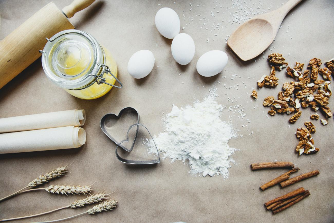 Photo of ingredients used in baking, laid out on a counter. A wooden rolling pin and a roll of parchment, along with a few sheafs of wheat are on the left of the image, while a small mound of flour is next to 2 heart-shaped cookie cutters in the center. Four eggs are nearby, as well as walnuts and stick cinnamon.