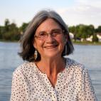 Photo of author Susan Casey. She is outdoors, and a lake with distant shore full of green trees is behind her. She has shoulder-length salt and pepper colored straight hair that is parted in the middle. She wears eyeglasses, and has long dangling earrings. She is smiling. Her blouse is white with small dark polka dots.