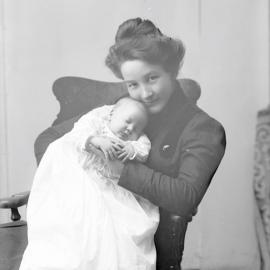 Black and white historical photo of a young woman holding a sleeping baby wearing a white gown. She is wearing black and staring at the camera with the child near her face.
