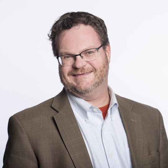 Photo of a man wearing a tan colored suit jacket over a light blue and white vertical striped button collar shirt which has the top button undone. He is not wearing a necktie, and the neckline of a reddish t-shirt is just visible. He has short, slightly curly brown hair and close-cut brown moustache and beard. He wears rectangular-shaped eyewear that has a black metal frame on the top half of the lenses.